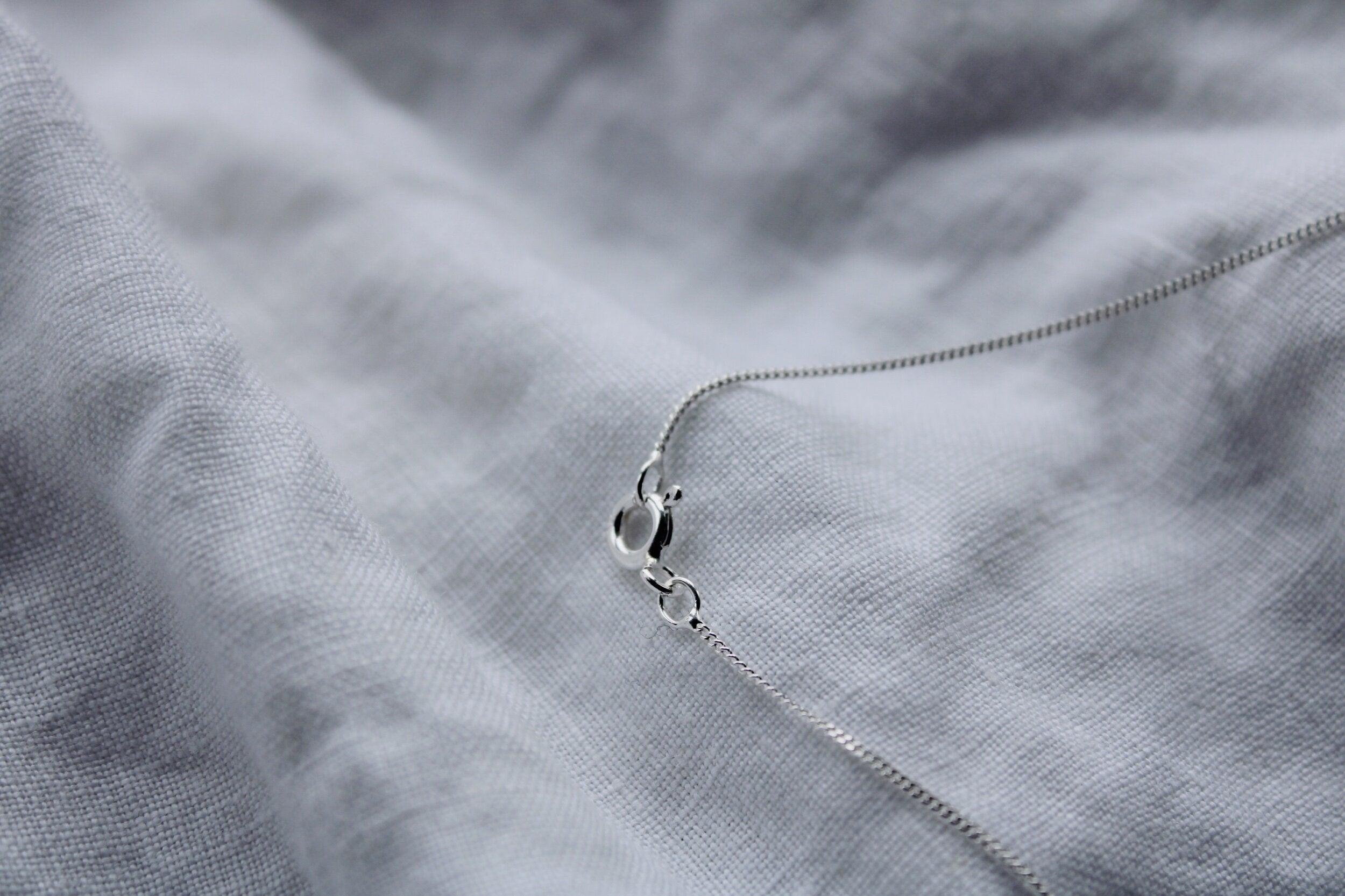 The Silver chain, crafted from delicate sterling silver with a small clasp, rests on soft light gray fabric, creating an elegant contrast in texture and color—an ideal choice for lovers of fine jewellery.