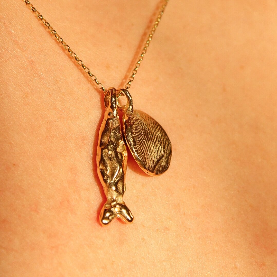 The Pescare necklace features a gold fish pendant and an oval charm with fingerprint texture, both elegantly displayed against bare skin.