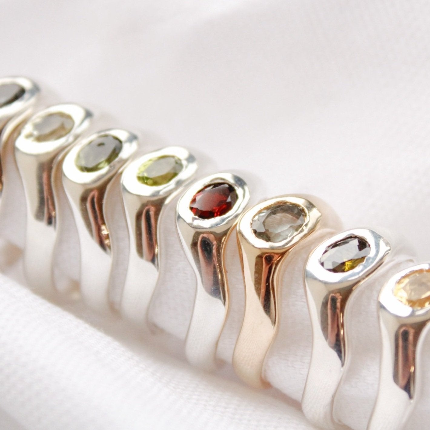 A row of The Silk rings in sterling silver and gold, featuring oval gemstones in assorted colors, displayed on a white fabric background.