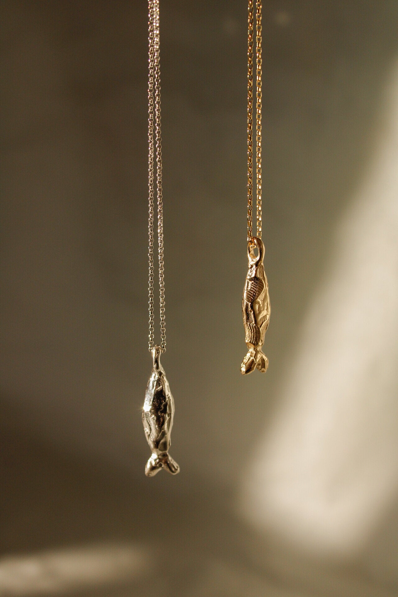 Two Pescare necklaces hang side by side: one with a sterling silver chain and seal pendant, the other with a gold chain and fish pendant, both set against a softly lit neutral background.