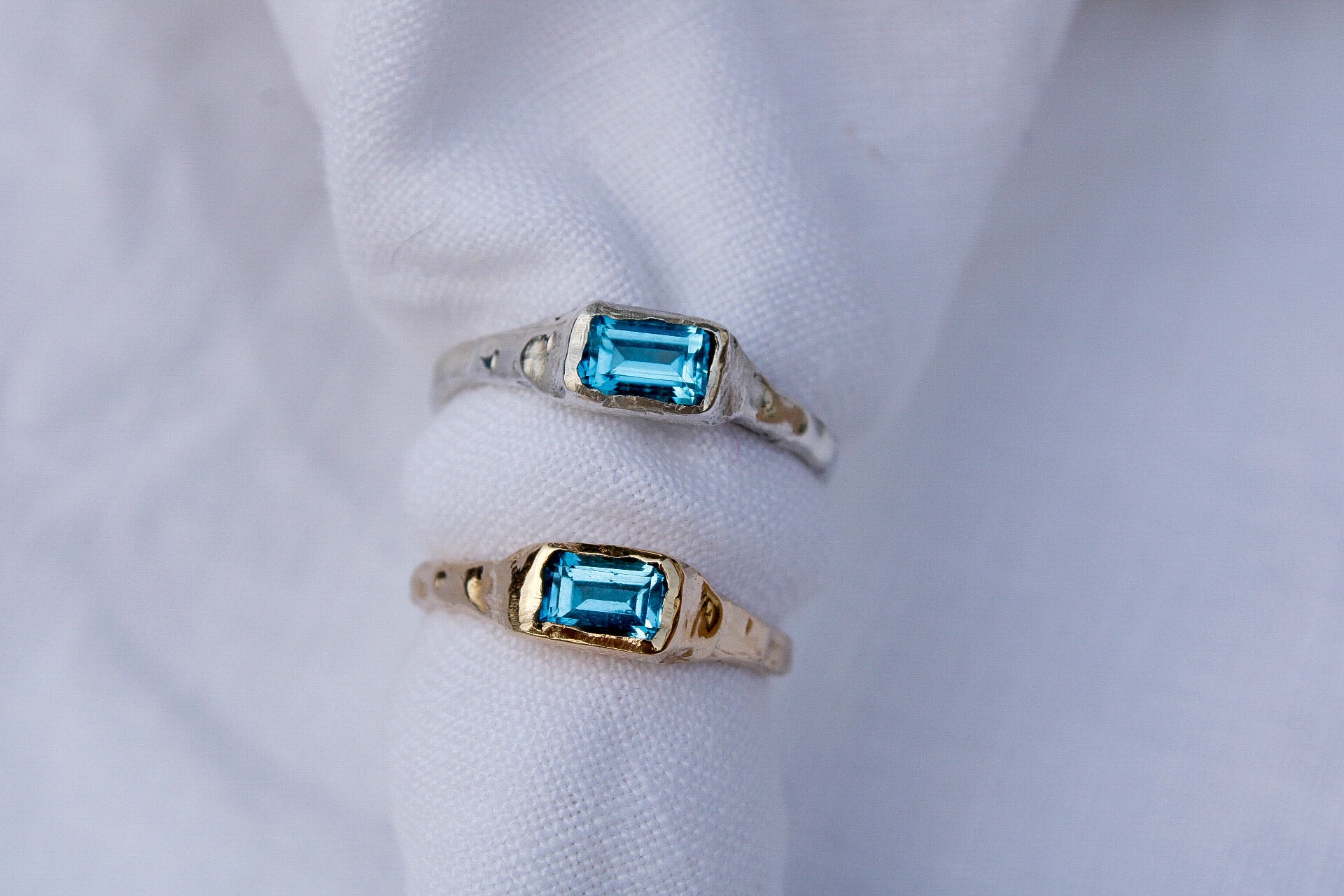 Two Pool rings with rectangular blue gemstones—one sterling silver, the other gold—are displayed on a white fabric background.