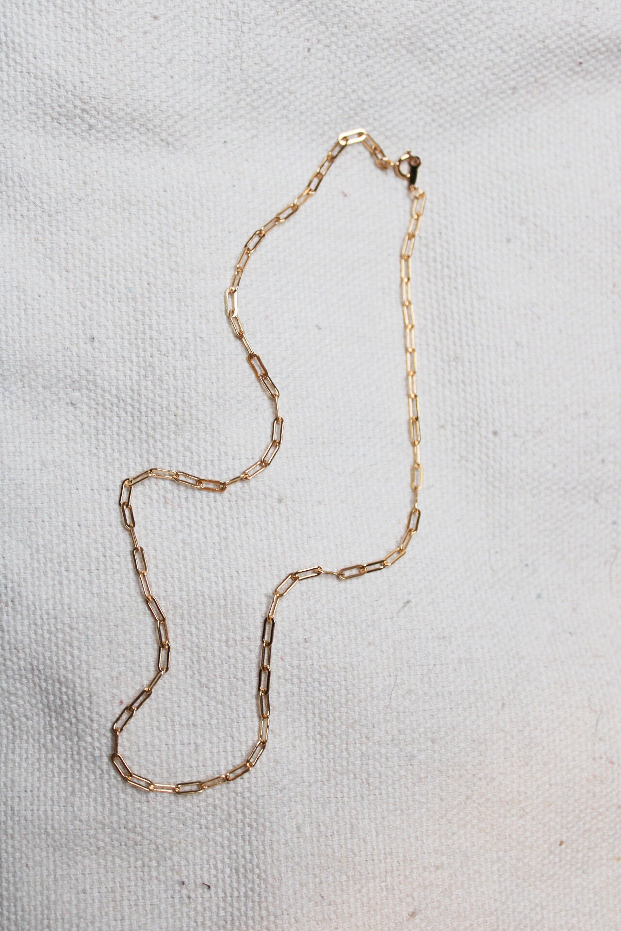 The Gold linked chain, featuring delicate rectangular links, is displayed on a textured off-white background—an elegant choice for everyday wear.