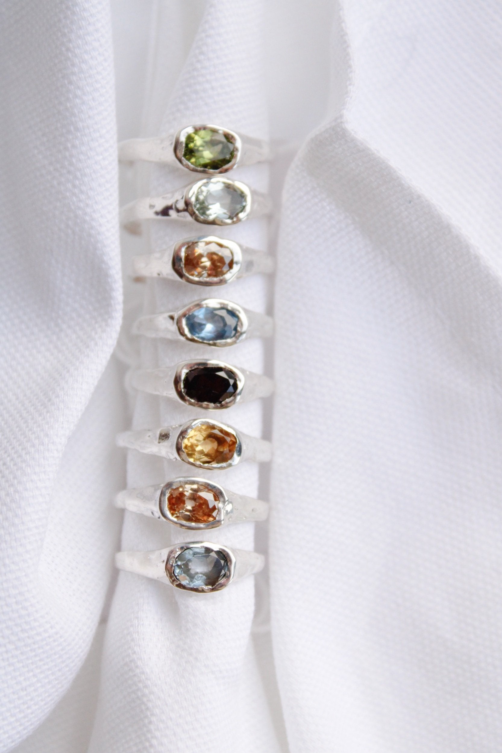 Seven “Florence” sterling silver rings, each with an oval gemstone in shades of green, yellow, blue, and brown, are displayed upright on textured white fabric, highlighting the distinctive charm of these statement rings.