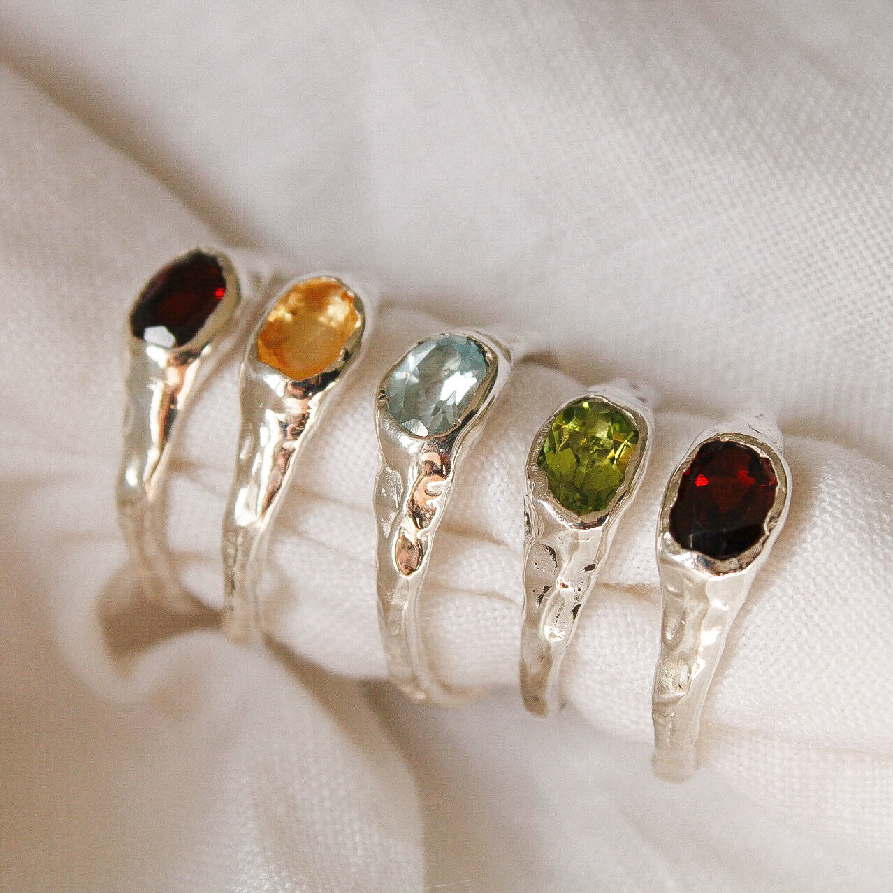 The Florence ring collection features five sterling silver rings with textured bands, each showcasing an oval gemstone in red, yellow, light blue, green, and deep red on a white fabric background.