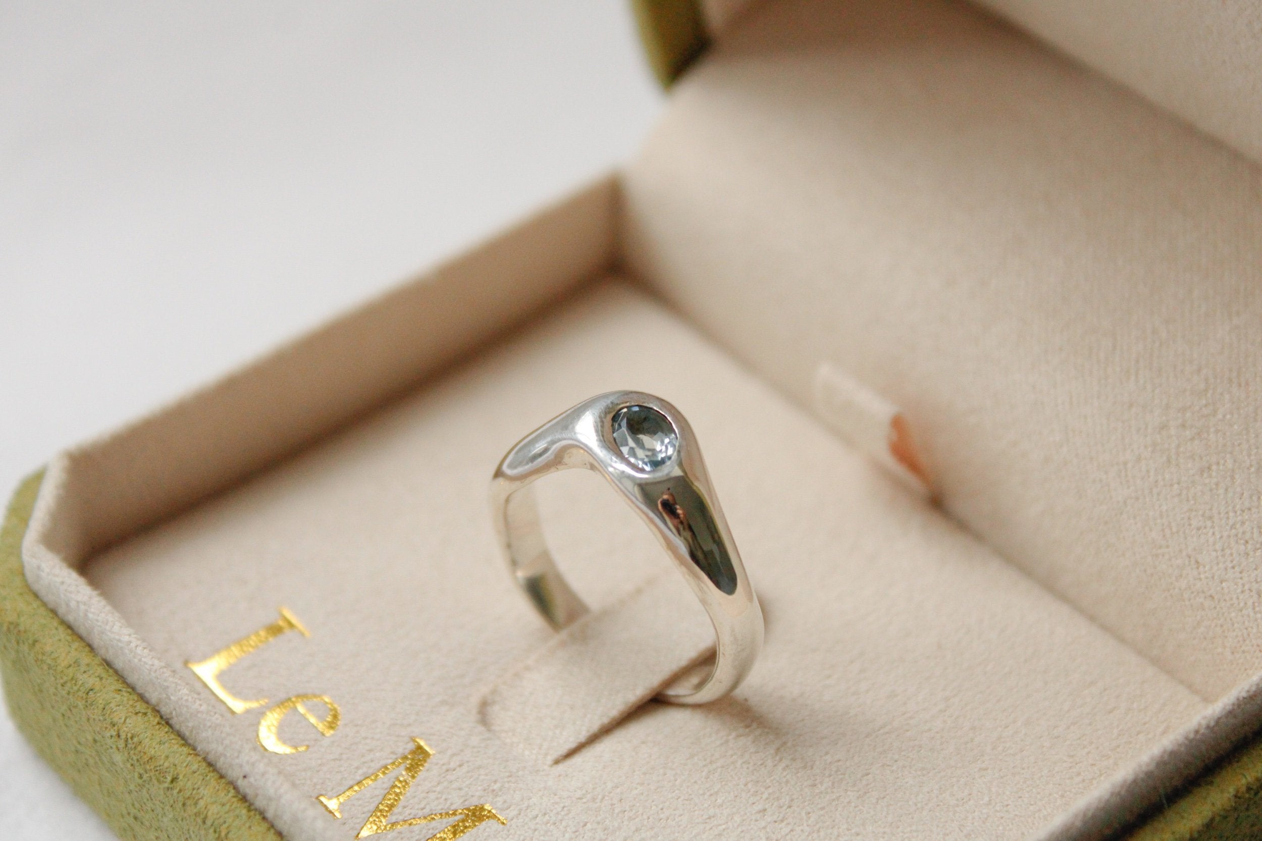 The Silk ring: a sterling silver band with a clear gemstone in a minimalist setting, presented in a beige velvet ring box featuring partial gold text inside.