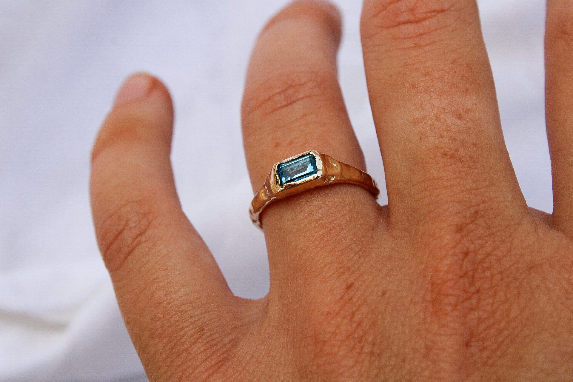 A hand displays the Pool ring—a bespoke gold band set with a rectangular blue gemstone—against a white background.