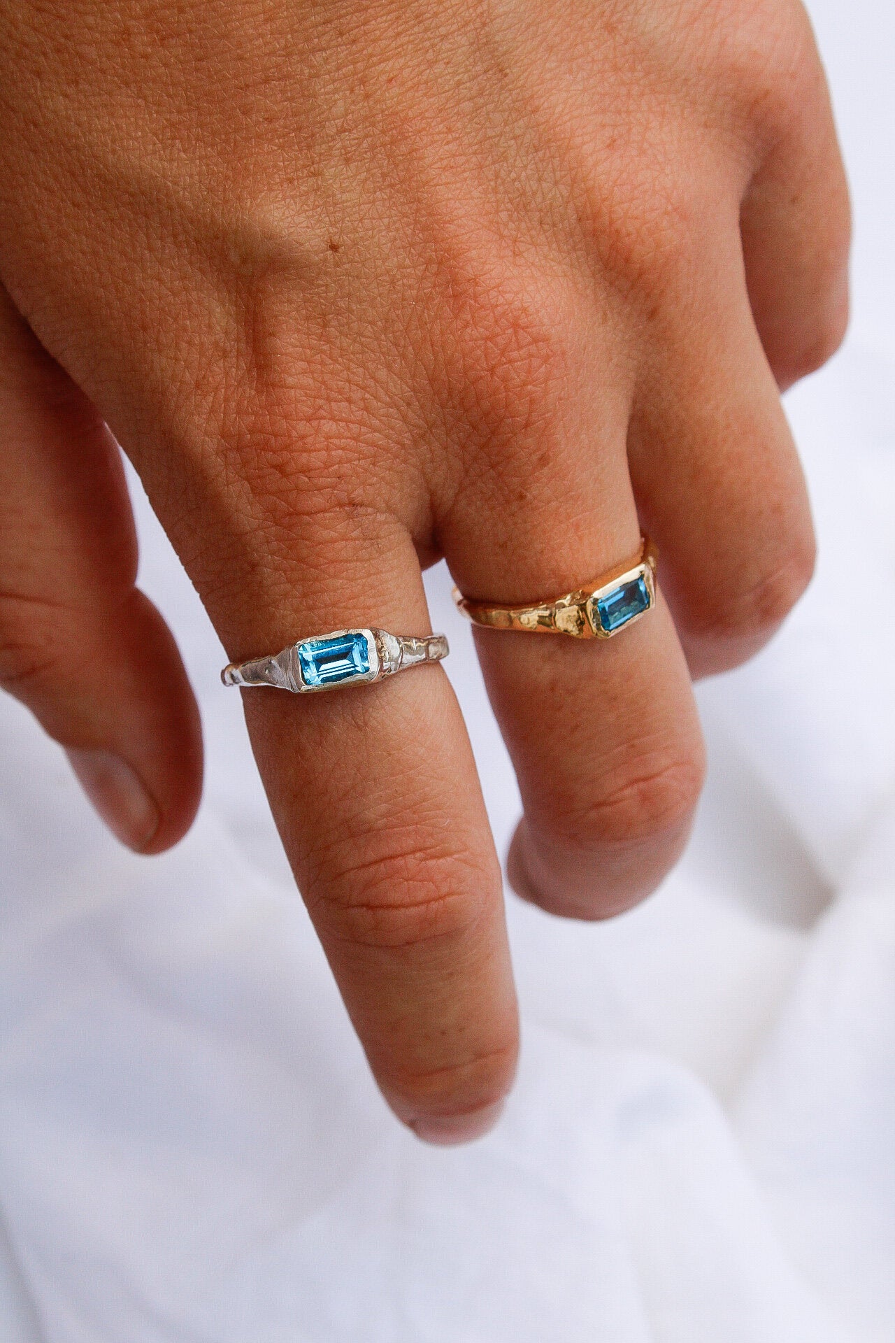 A hand wears a handmade silver ring with a rectangular blue gemstone on the middle finger and the Pool ring in gold on the ring finger, set against a white fabric background.