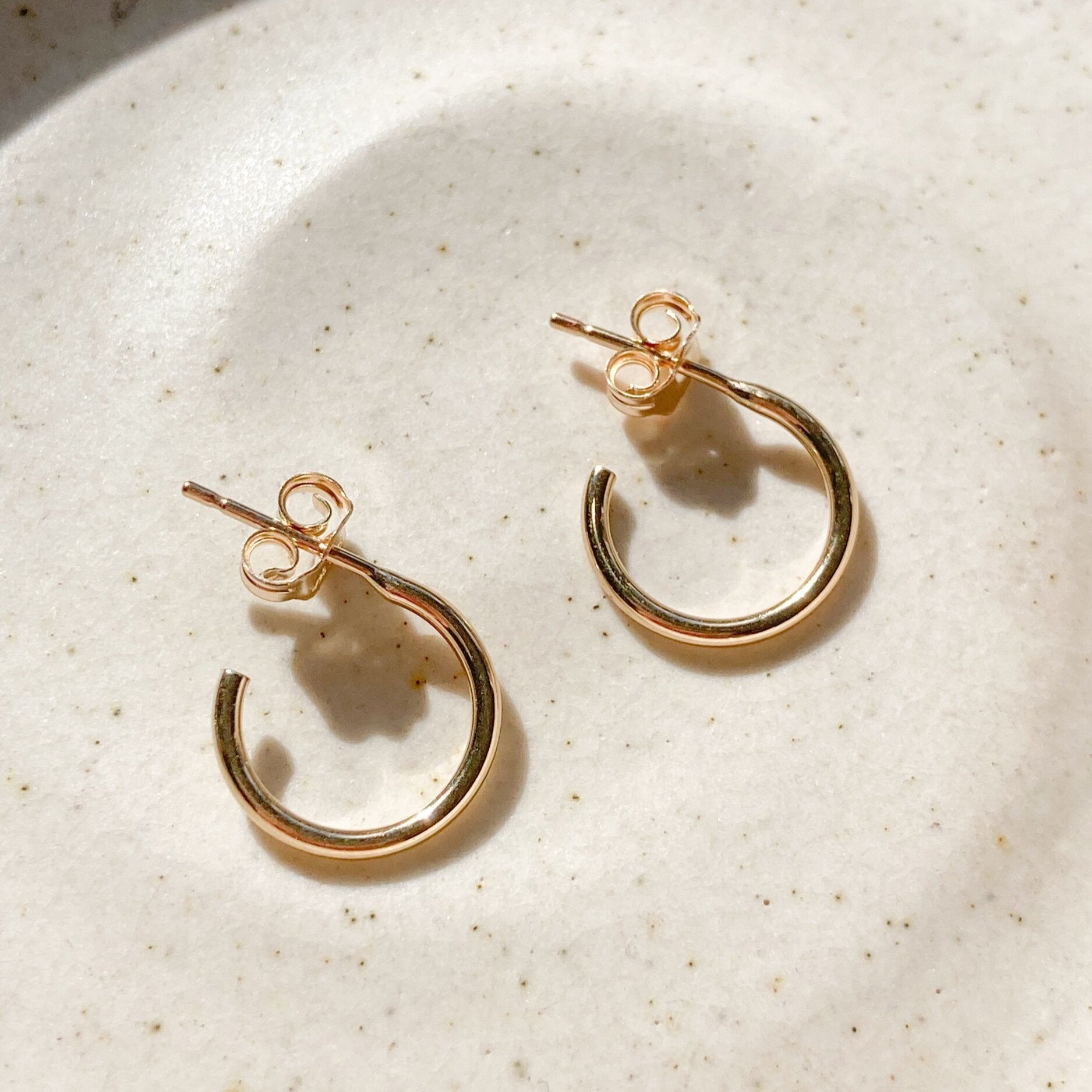 Gabriella’s hoops are small gold-filled hoop earrings with butterfly backings, shown on a speckled cream ceramic surface in soft natural light—perfect waterproof earrings for daily wear.