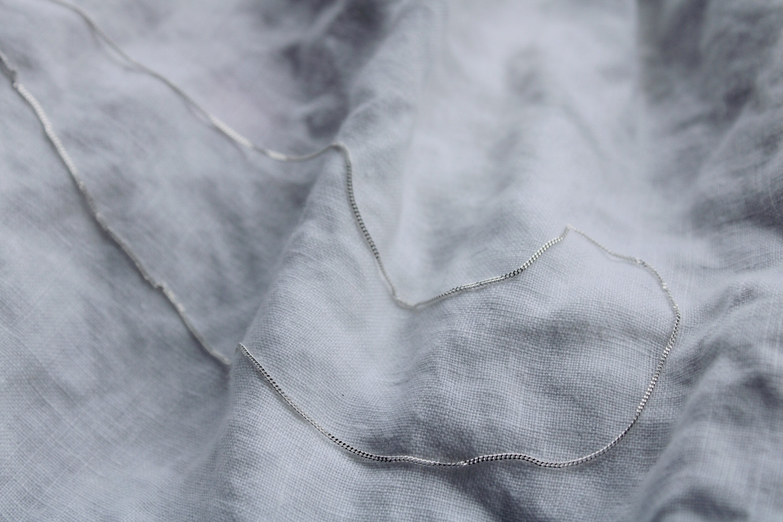 The Silver chain, featuring a polished sterling silver finish, lies on softly crumpled light gray fabric, creating gentle curves and shadows—a subtle showcase of elegant jewellery.