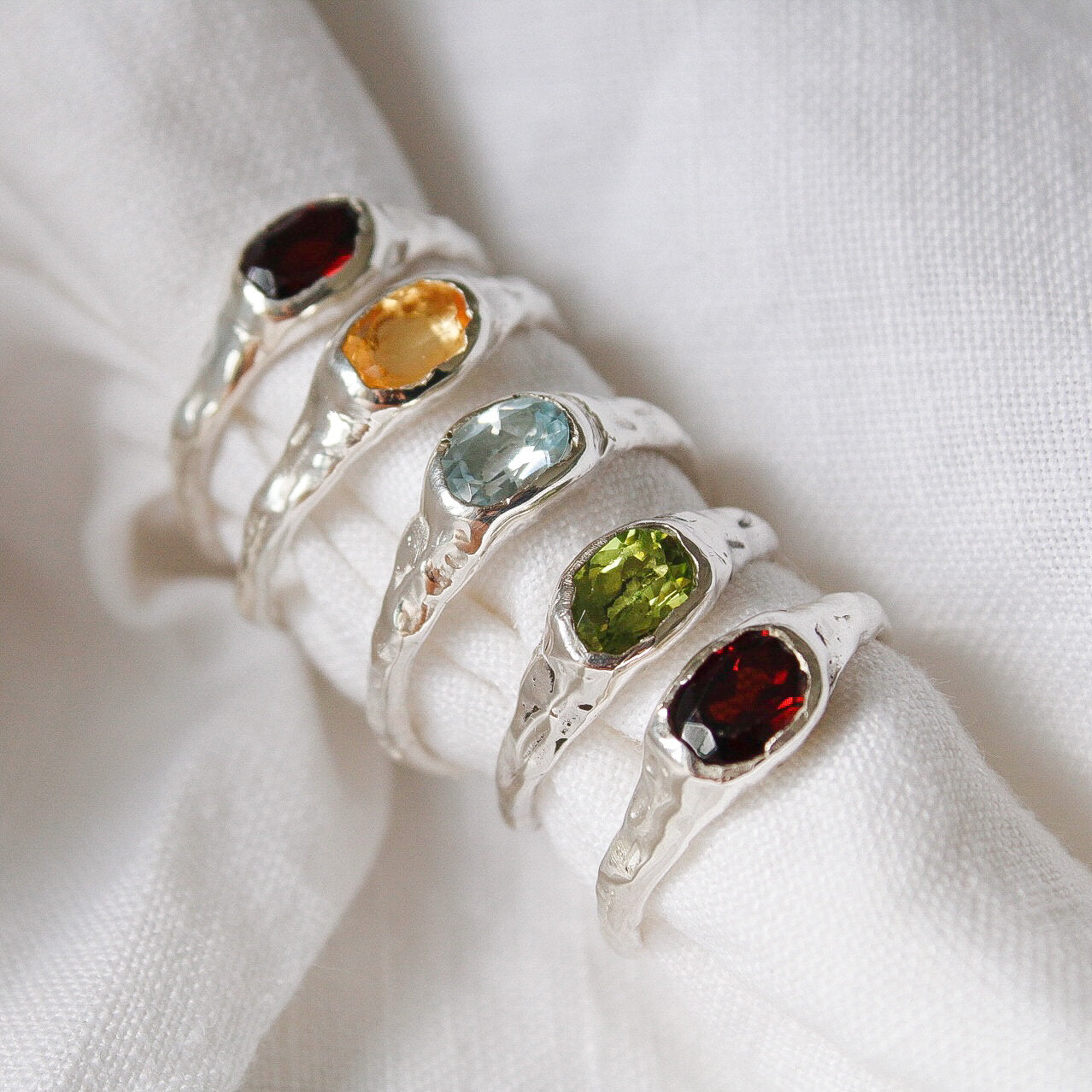 Five Florence rings in sterling silver with red, yellow, light blue, green, and dark red gemstones are displayed on white fabric. Each unique bespoke ring features a textured band for a standout look.