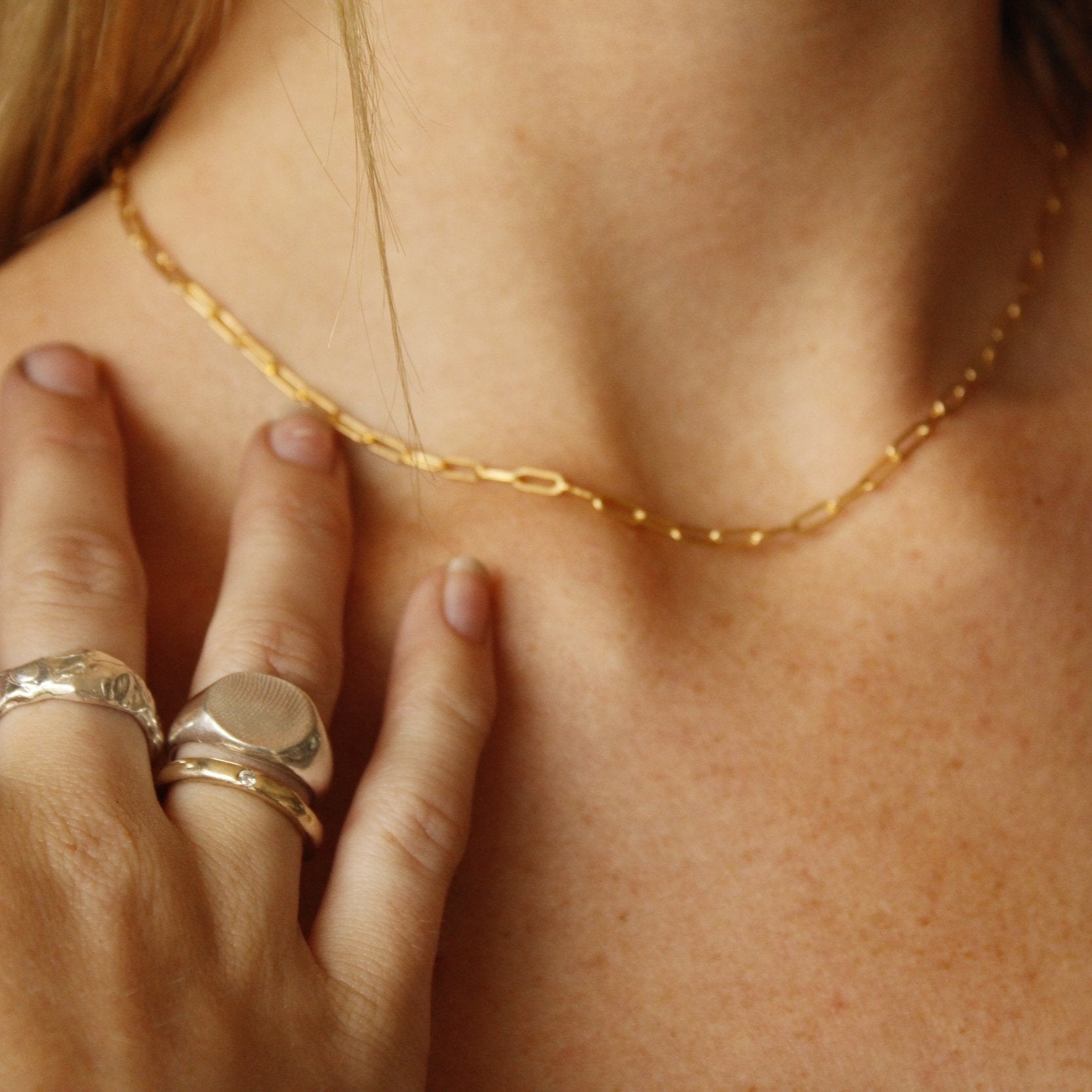 A close-up of a hand with silver rings touching a collarbone, wearing the Gold linked chain. The softly blurred background highlights this everyday necklace, emphasizing the jewelry and warm skin tones.