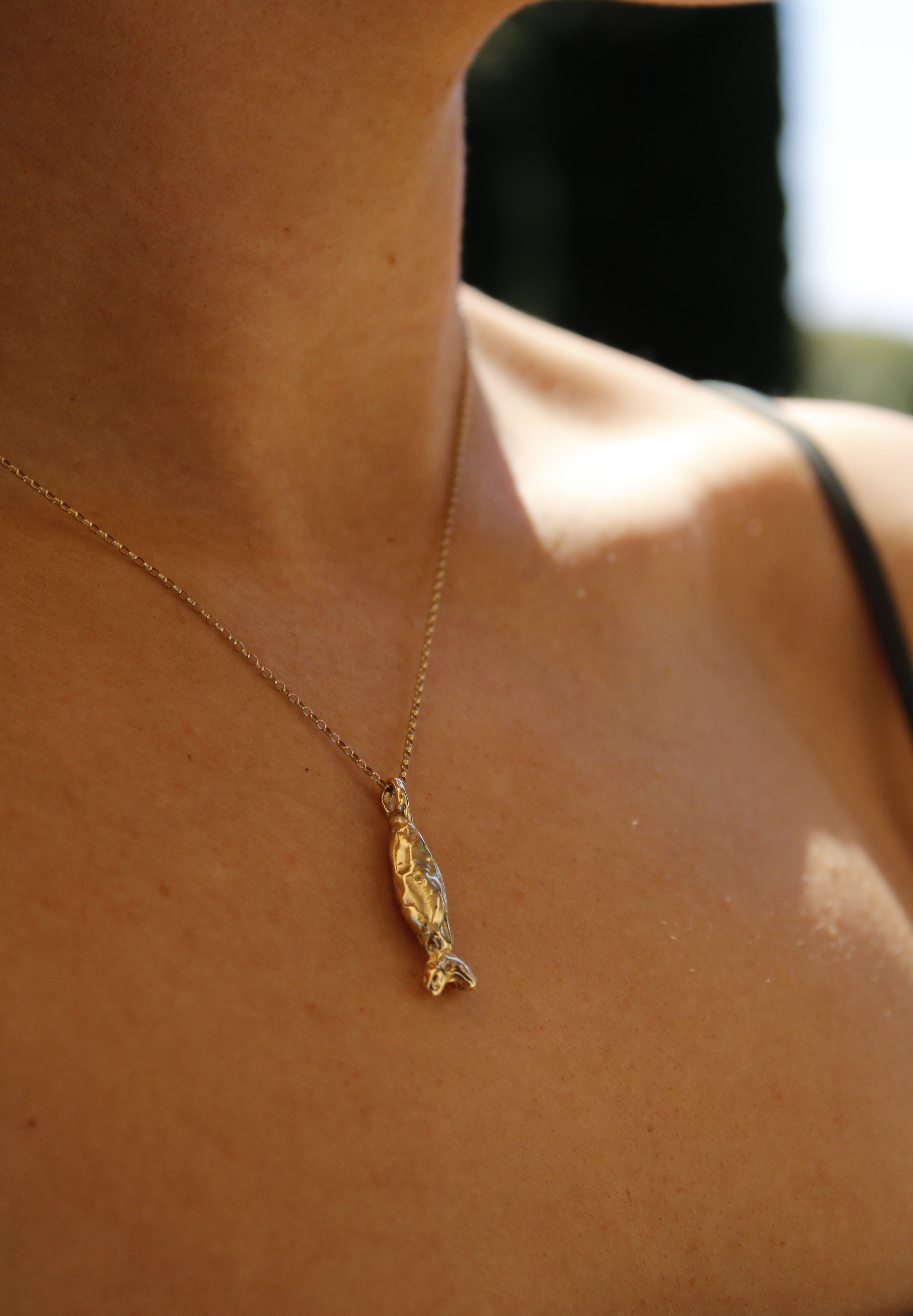 A close-up of a person wearing the Pescare necklace—a gold fish pendant—against bare skin and a black strap, softly illuminated by sunlight.
