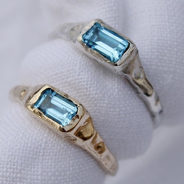 Two Pool rings with rectangular blue gemstones are displayed side by side—one with a gold band and the other a handcrafted recycled sterling silver band—both set against a white textured background.
