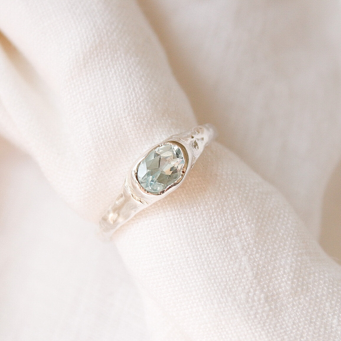 The Florence ring, a delicate sterling silver piece featuring an oval light blue gemstone, is displayed on a soft white fabric background.