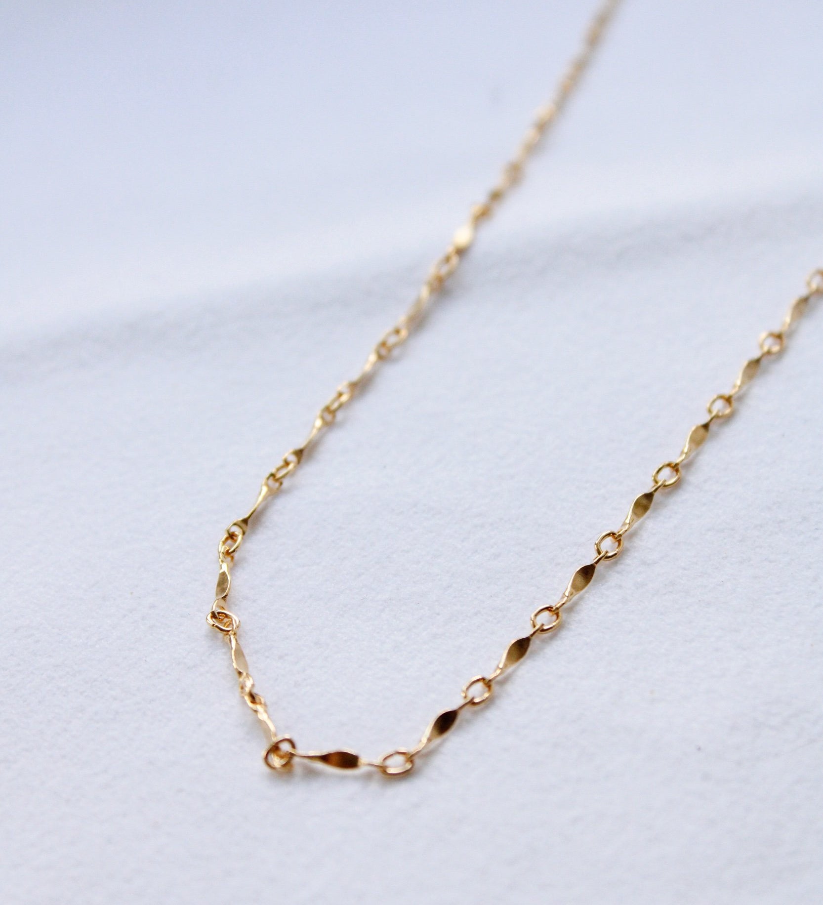 The Twisted Link Necklace features a gold filled chain with small oval and twisted links. Displayed on a white textured surface, this waterproof, minimalist necklace is perfect for everyday elegance.