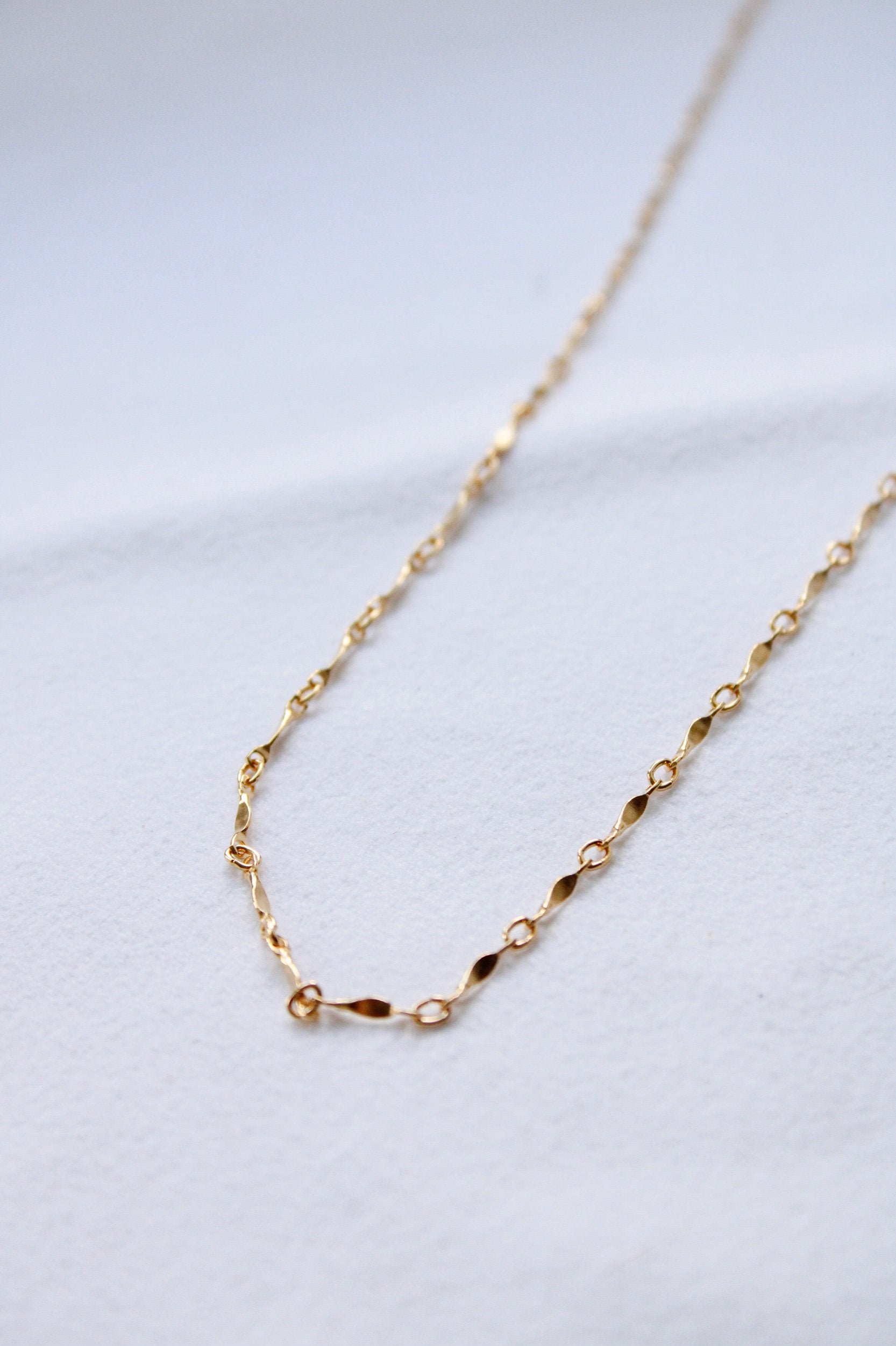The Twisted Link Necklace features a gold filled chain with small oval and twisted links. Displayed on a white textured surface, this waterproof, minimalist necklace is perfect for everyday elegance.