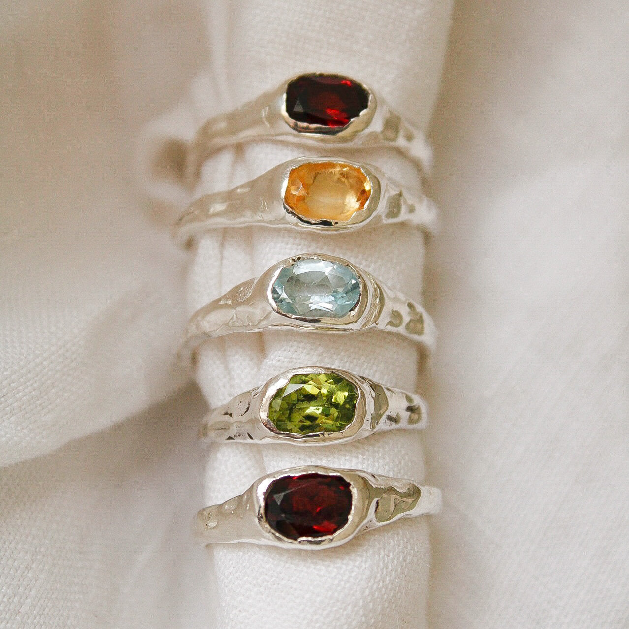 Five sterling silver Florence rings, each with a distinct gemstone (red, orange, blue, green, and dark red), are stacked vertically on a white cloth. Each Florence ring is a unique statement piece for any occasion.