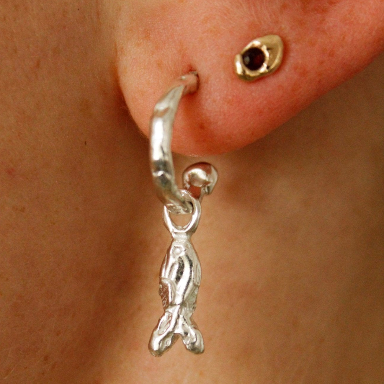 A close-up of an ear wearing two earrings: a gold stud with a dark red gem in the upper piercing and Charm hoops—crafted from recycled sterling silver with a fish-shaped dangle—in the lower piercing.