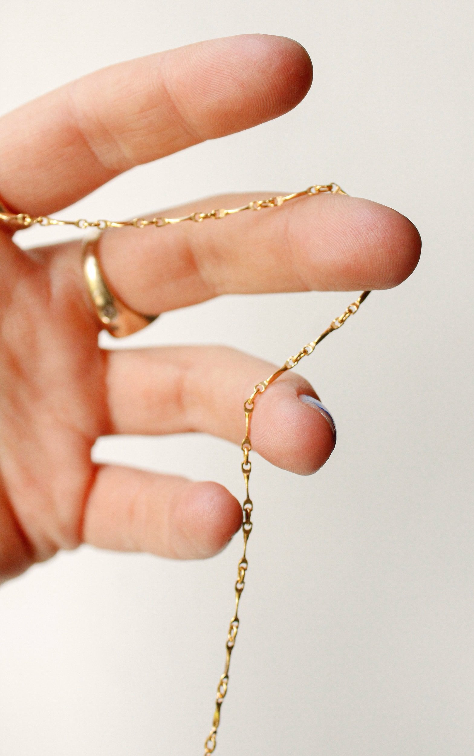 A close-up of a hand holding the Twisted link necklace—a delicate gold filled chain—while a gold ring glimmers on the ring finger. The plain, light background highlights the minimalist design of this elegant necklace.
