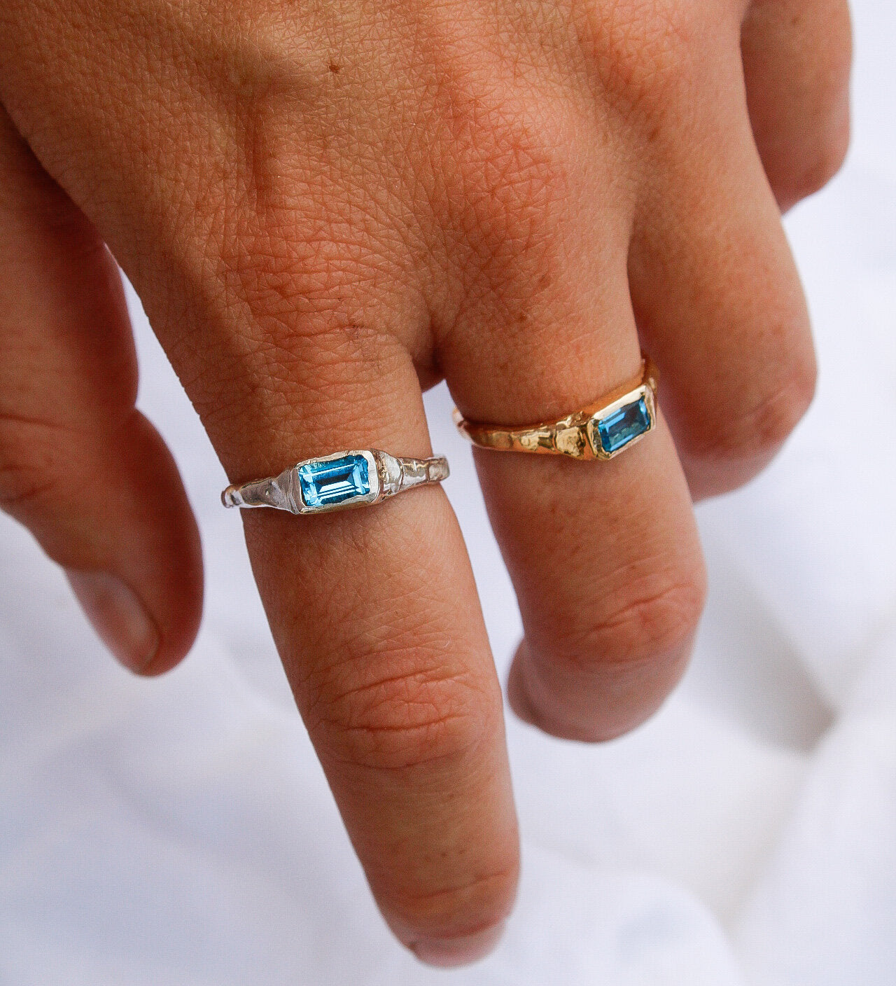 A hand wears a handmade silver ring with a rectangular blue gemstone on the middle finger and the Pool ring in gold on the ring finger, set against a white fabric background.