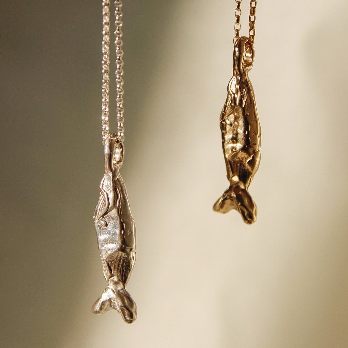 The Pescare necklace features two metallic fish pendants—one sterling silver, one gold—hanging from delicate chains and showcased against a softly blurred background.
