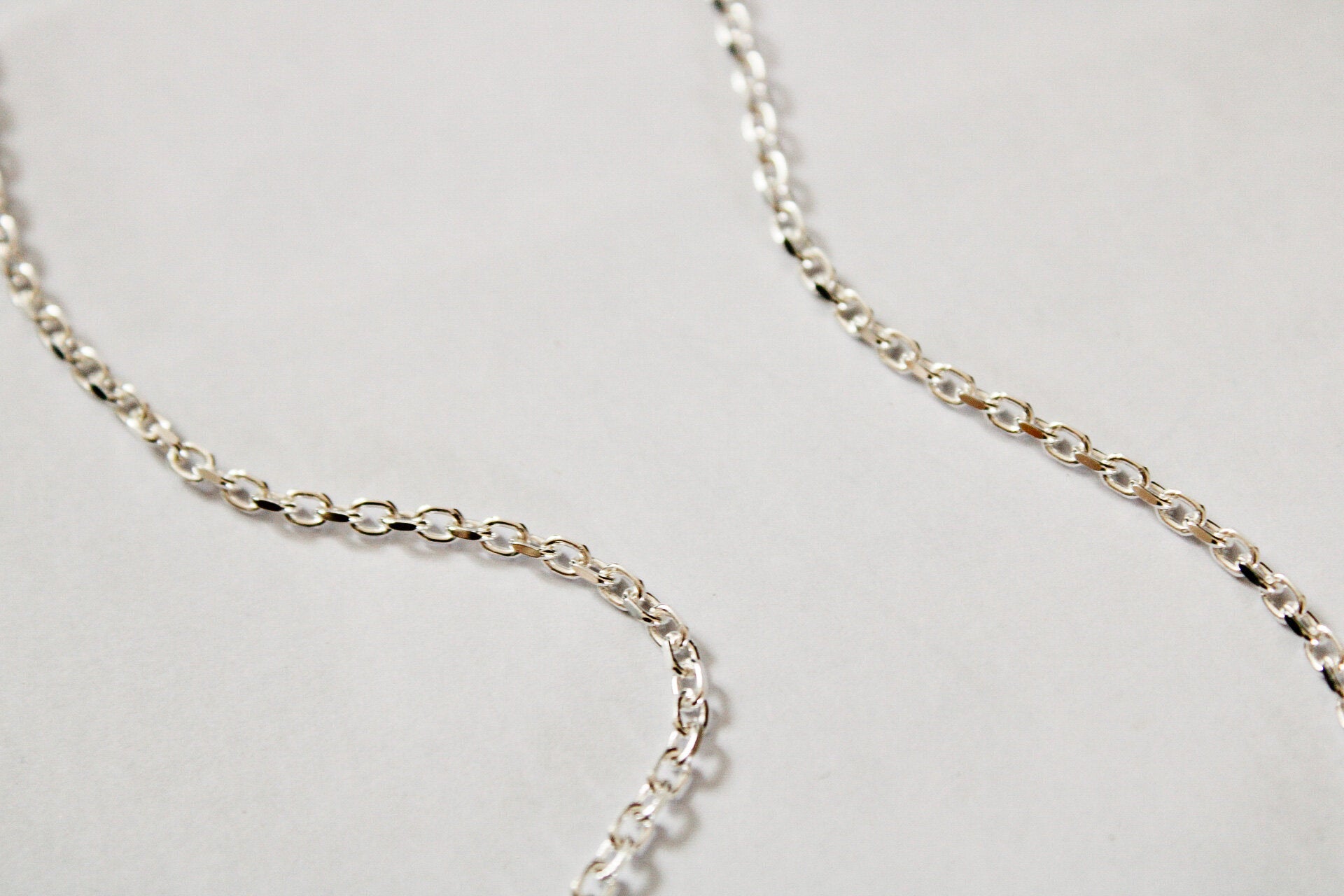 A close-up of the Mens Chain, a delicate sterling silver Belcher chain with small oval links, gracefully curves and loops on a plain light background.