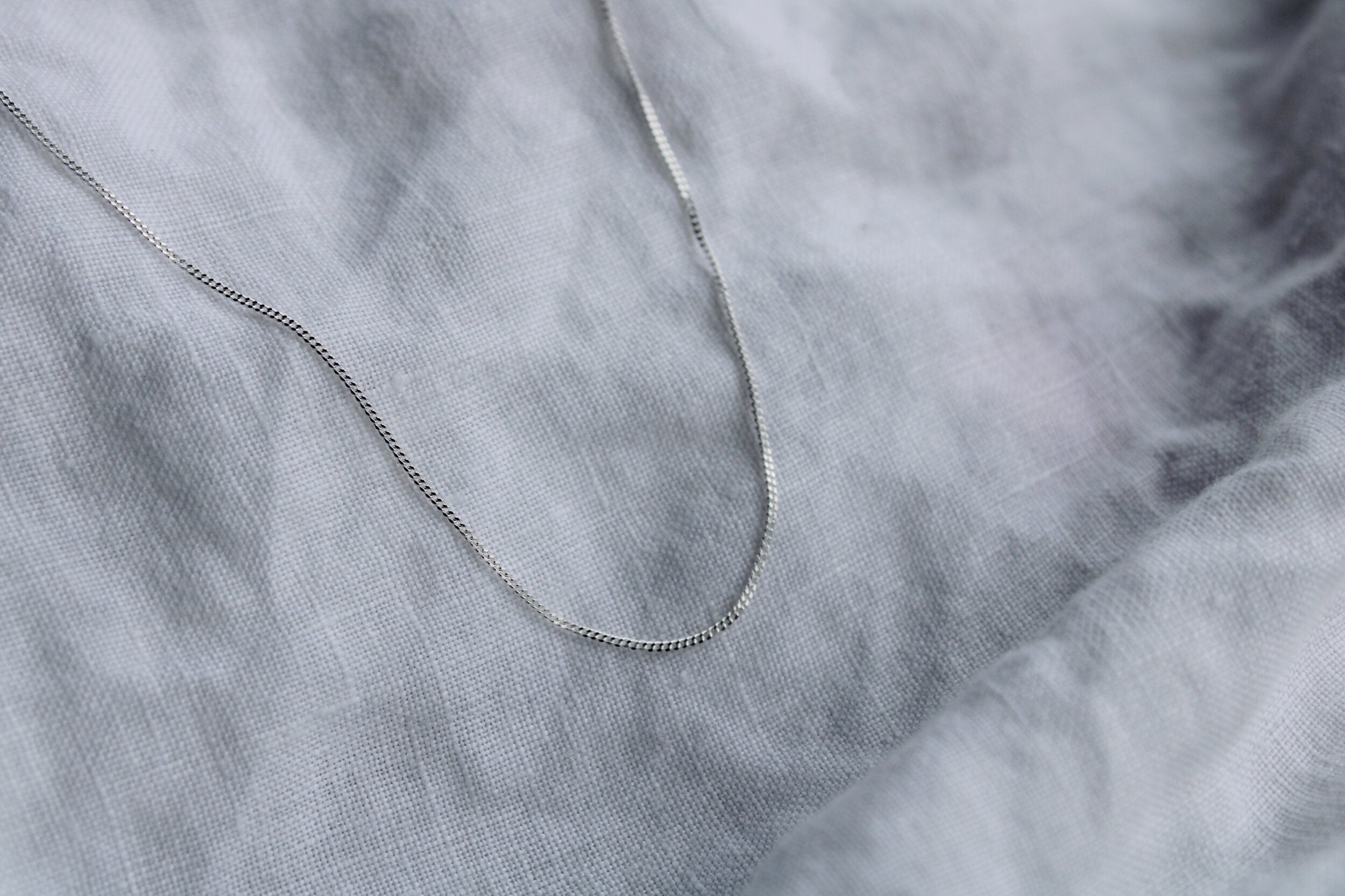 The Silver chain is a delicate sterling silver necklace displayed on soft, white, slightly wrinkled fabric—perfect for fine jewellery lovers.