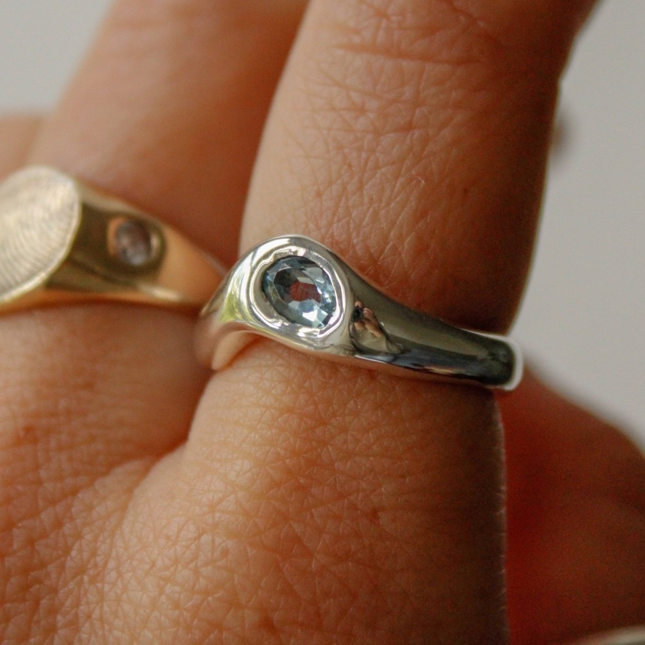 A close-up of a hand wearing The Silk ring, a sterling silver piece featuring a blue gemstone and exquisite craftsmanship, alongside a gold ring with an engraved design and a small clear stone.