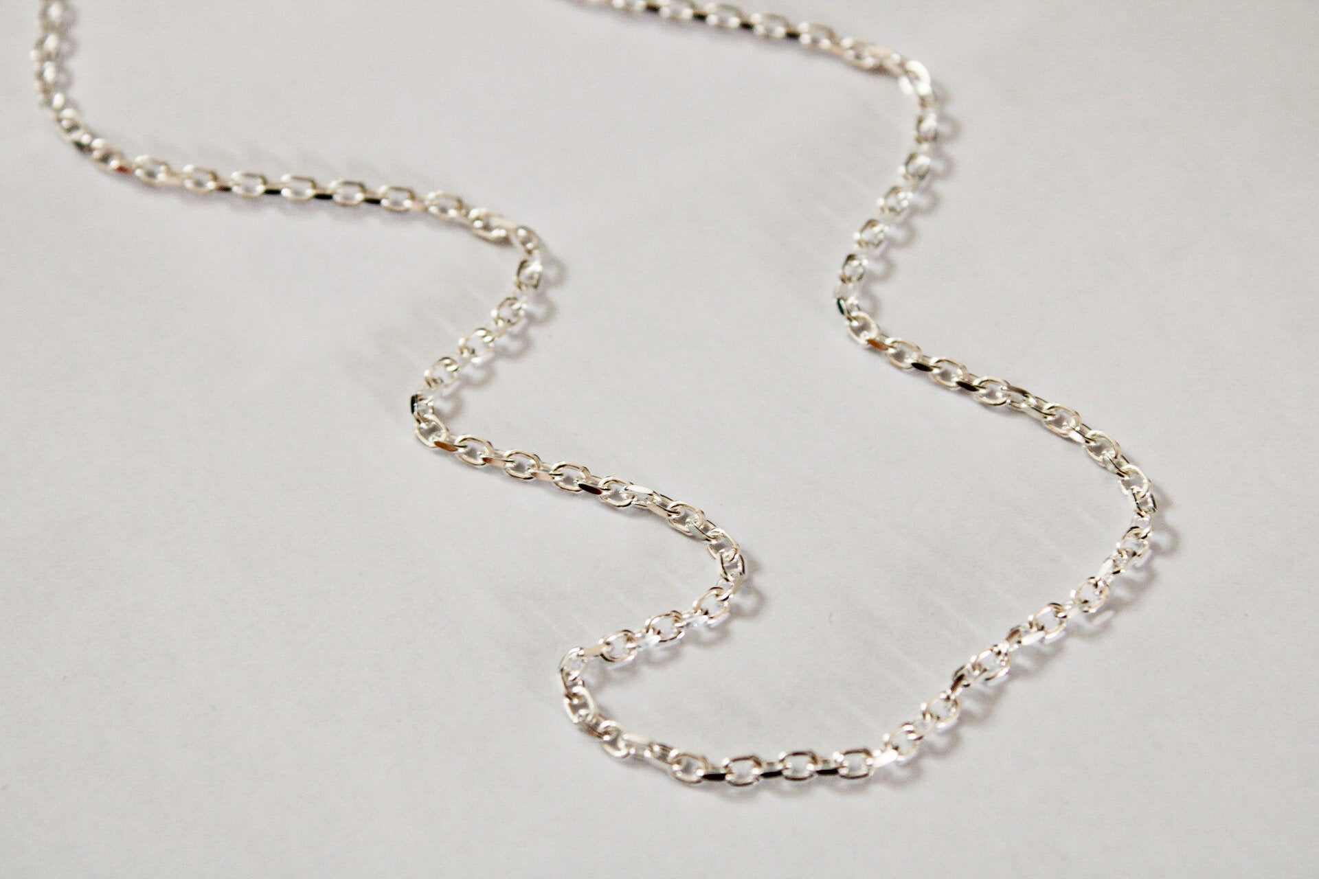 A Mens chain in sterling silver, featuring small oval Belcher links, is arranged in a loose, wavy shape on a plain white background.