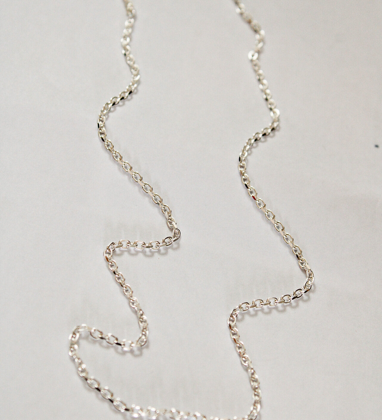 A Mens chain made of thin sterling silver is displayed in a loose, irregular shape on a plain, light-colored surface.