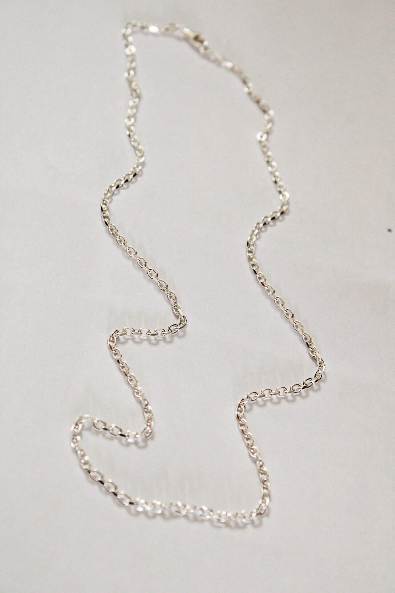 A Mens chain made of thin sterling silver is displayed in a loose, irregular shape on a plain, light-colored surface.