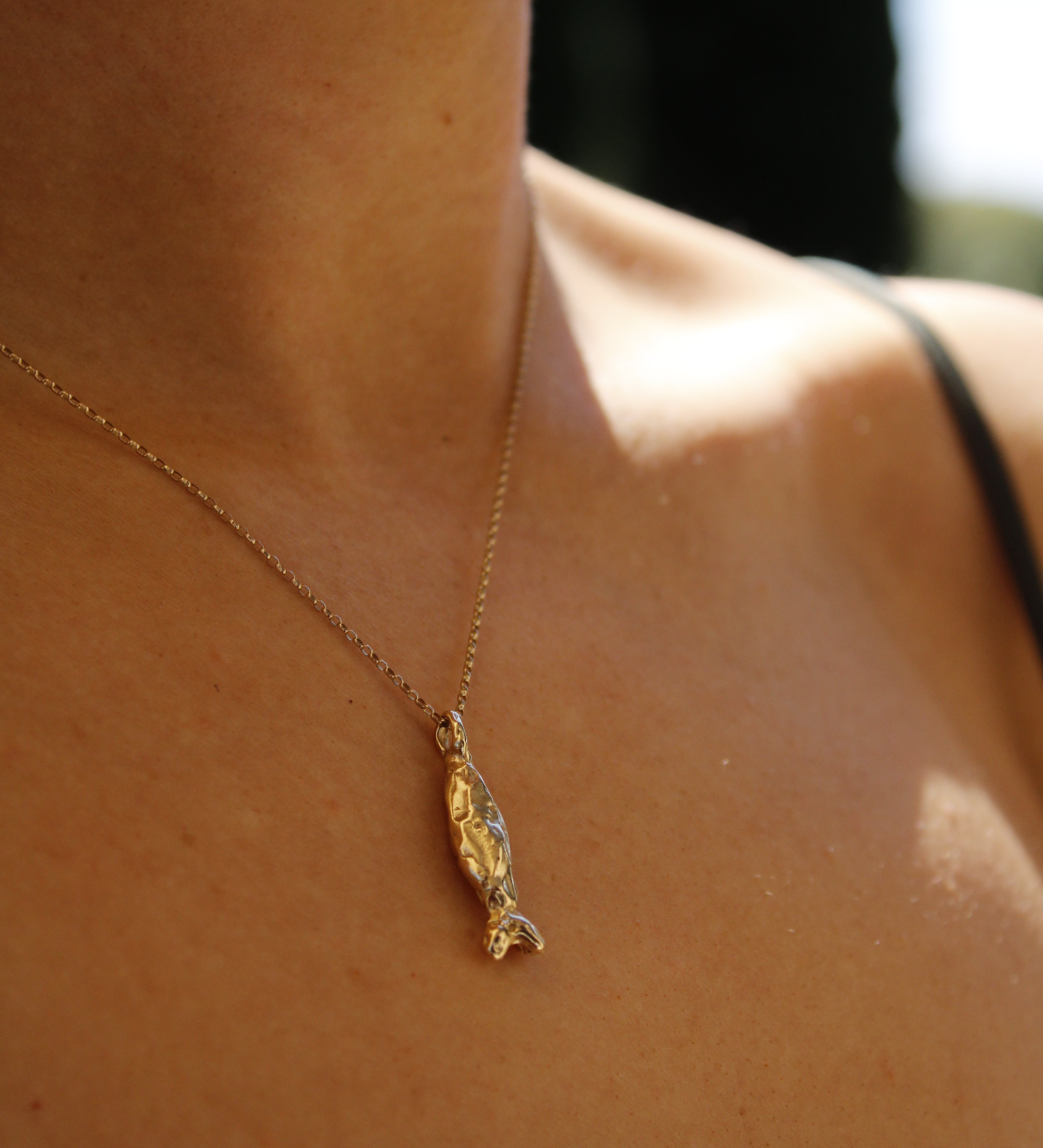 A close-up of a person wearing the Pescare necklace—a gold fish pendant—against bare skin and a black strap, softly illuminated by sunlight.